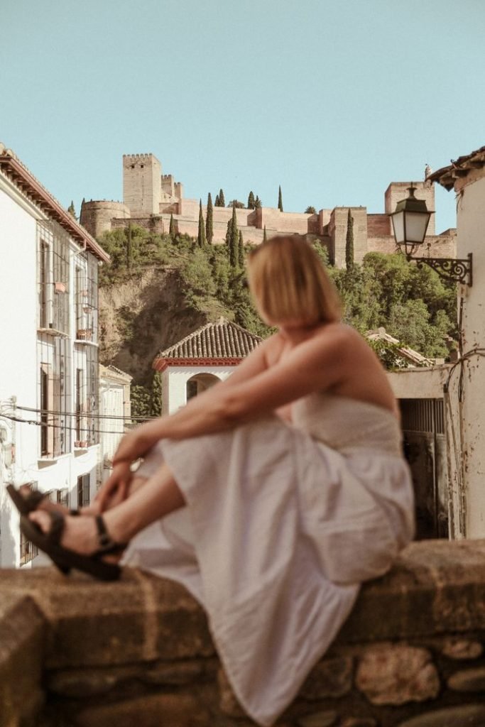 A woman in a white dress sits on a stone wall, overlooking the Alhambra in Spain.